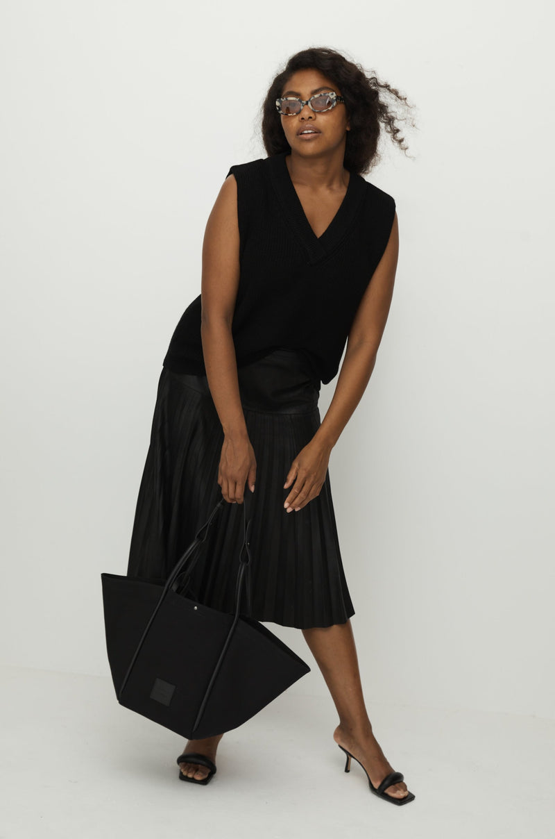 Women in black top and skirt holding a black canvas tote bucket bag, with black leather rolled leather handles, silver button closure at the top and small square logo of WEST14TH and X NIHILO.