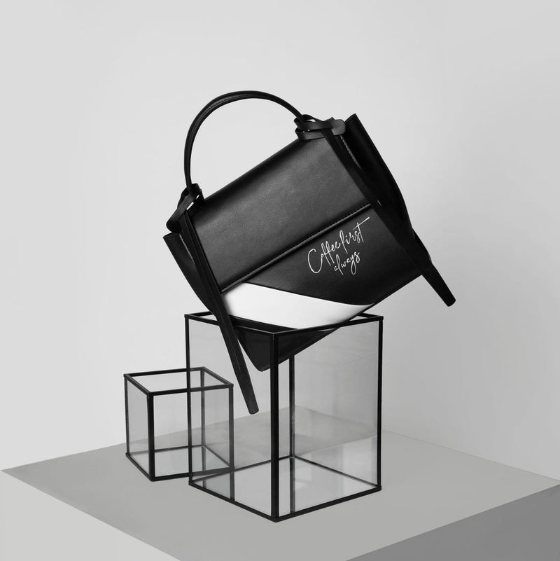 Rectangle genuine black and white cow nappa leather luxury work bag and handbag with black leather tassels, front flap and handle positioned on an angle on top of a clear rectangular prism. Customised text "Coffee first always" written on the bag.