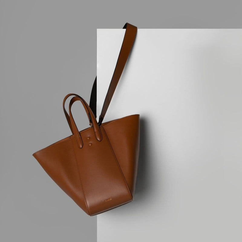 X NIHILO Eight in tan bag, fashion bag with single snap button top closure, soft gold hardware, and adjustable shoulder straps, luxury cow nappa leather handbag, genuine leather bag shown hanging off the edge of a door by its strap.