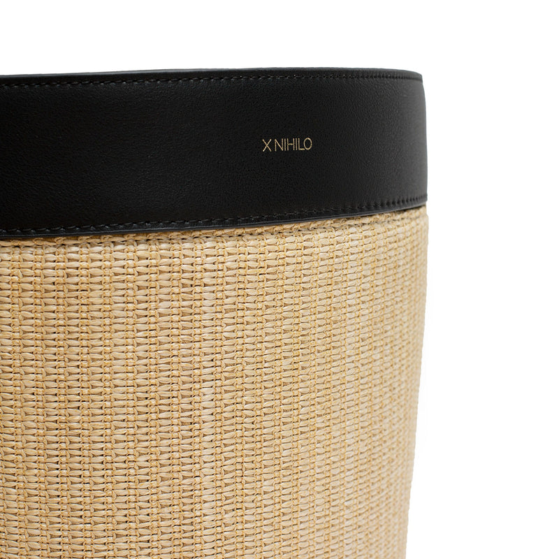 Close up of raffia picnic basket with black leather top on the side and detachable leather handle on the back. Gold logo embossed on the top with soft gold hardware on the sides. Inside is lined with insulated material and a top zip closure.